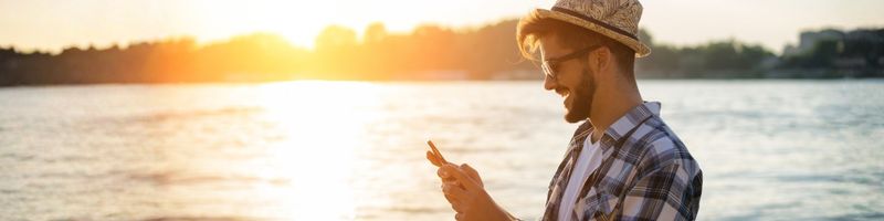 Photograph of a young man standing alongside a body of water at sunset, looking at his mobile phone and smiling