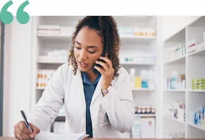 Pharmacist in lab coat writes while on phone in a pharmacy.
