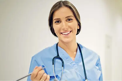Smiling healthcare worker in scrubs with stethoscope and pen indoors.