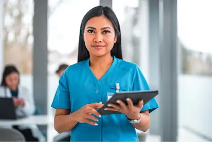 Healthcare professional in blue scrubs using a digital tablet in a bright clinical setting.