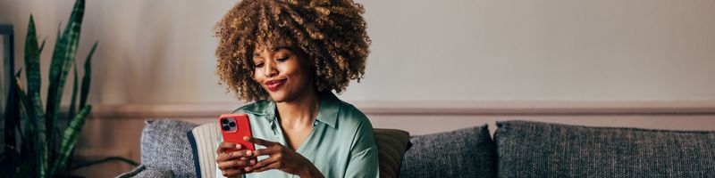 Photograph of a black woman in a collared shirt casually sitting on a couch smiling while she looks at her mobile phone