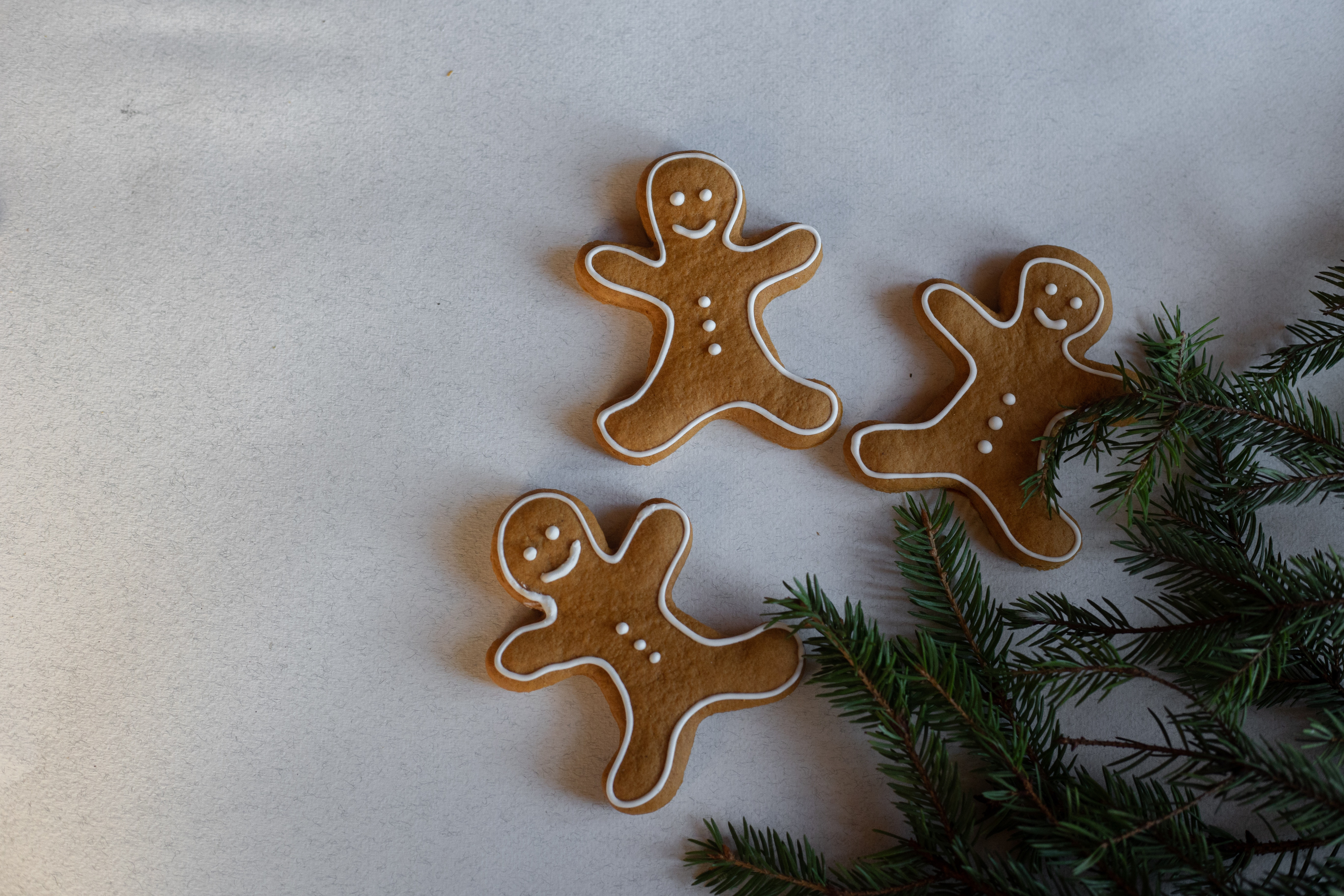 Three gingerbread cookies shaped like cartoon people, with thin white lines of icing tracing their outlines and forming their eyes, smiling mouth, and three shirt buttons. A few branches of a Christmas tree are just barely visible in the bottom right corner.