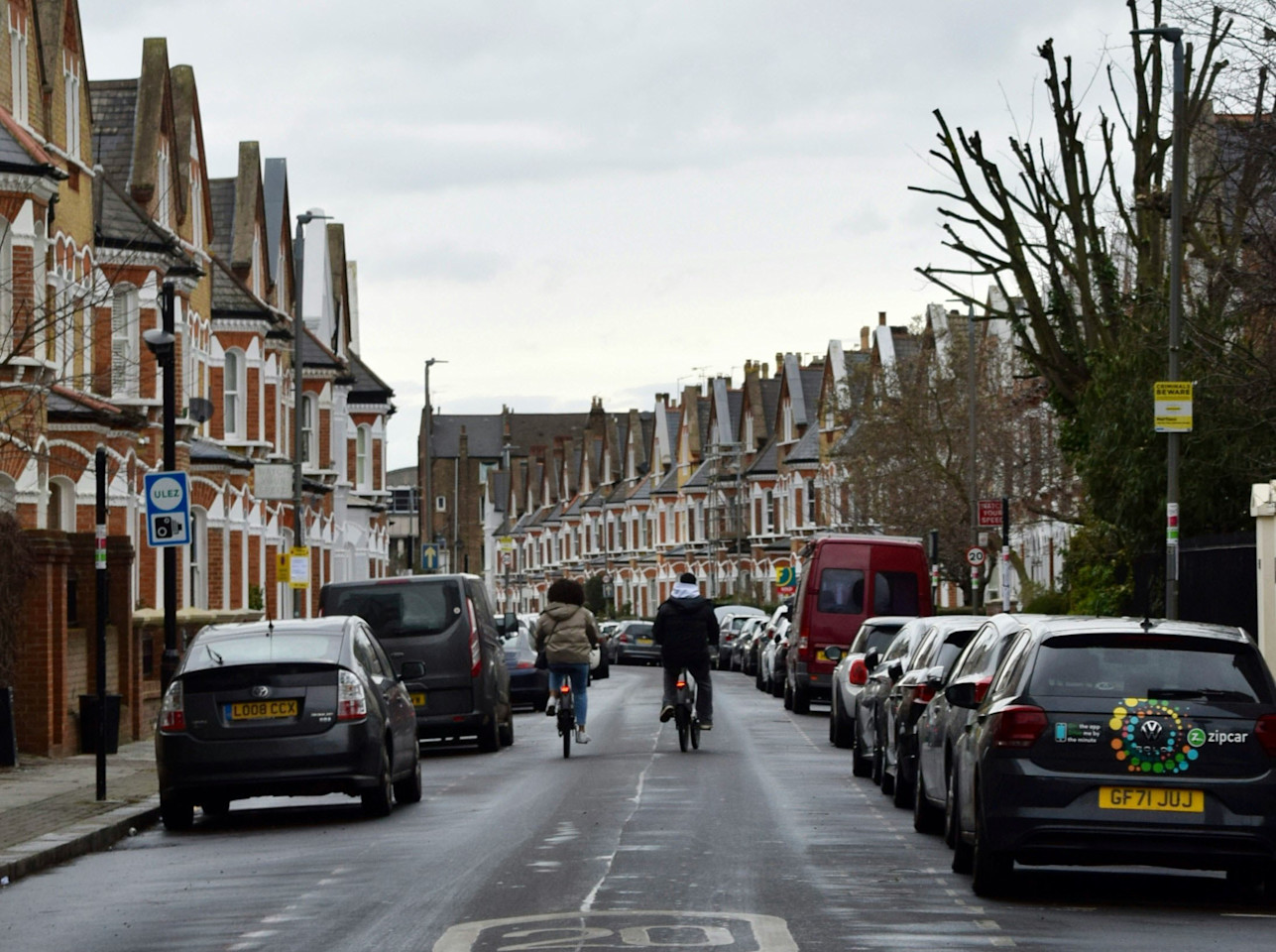 A photo looking down the road of a residential street in Britain with cars parked on either side and two cyclists riding down the road