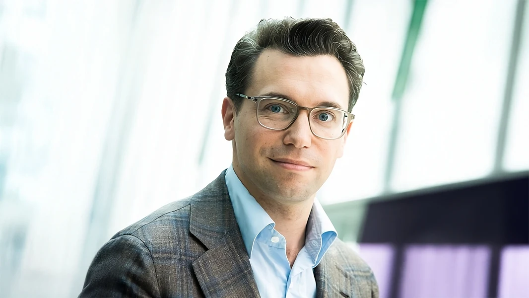 A professional portrait of Paul Fournier in business attire. He is wearing glasses, a checked blazer, and a light blue shirt. The background is modern and bright, with soft, blurred lines suggesting an office or conference room setting. The man looks friendly and confident at the camera.