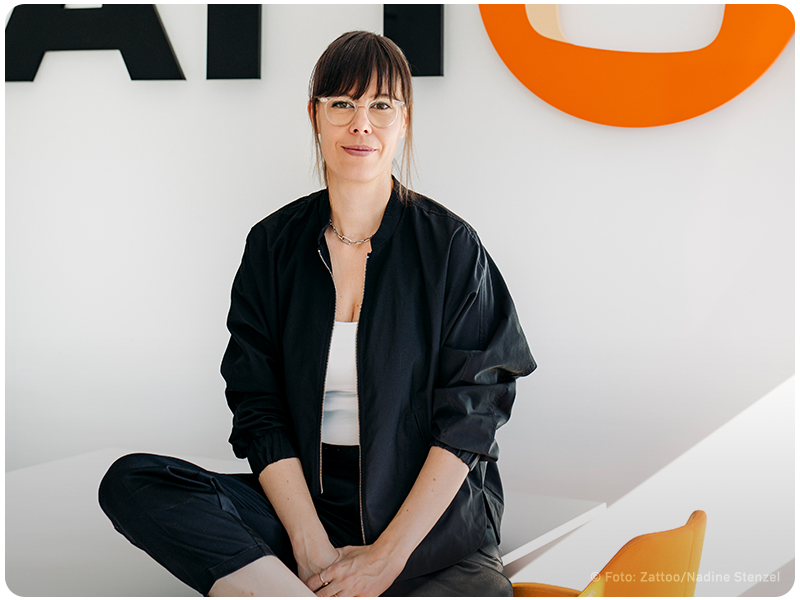 Nikoletta Tamas, in schwarzer Jacke und weißem Oberteil, sitzt entspannt vor dem Zattoo-Logo im Büro. Sie trägt eine Brille und lächelt leicht in die Kamera, mit dem großen Zattoo-Logo im Hintergrund.