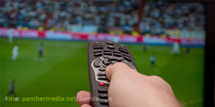 Eine Nahaufnahme einer Hand, die eine Fernbedienung hält und auf einen Fernseher zeigt, auf dem eine Sportübertragung mit einem Fußballspiel läuft. Im Hintergrund sind unscharfe Spieler auf dem Spielfeld und ein gefülltes Stadion zu sehen.