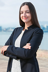 Kelly stands in a relaxed three-quarter pose, smiling, wearing a medium grey blazer over a striped shirt and dark jeans against a light grey background.