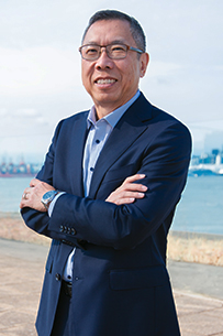 Phil stands in a relaxed three-quarter pose, smiling, wearing a purple polo shirt with dark jeans and a light belt against a light grey background.