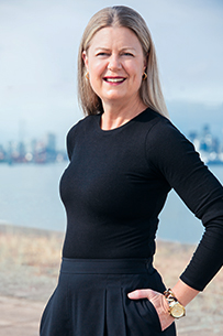 Jaci stands in a relaxed three-quarter pose, smiling, dressed in a dark cardigan sweater over a dark shirt with dark jeans on a light grey background.