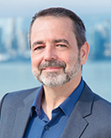 Jason smiles at the camera, wearing a blue collared shirt and dark grey suit against a light background. 