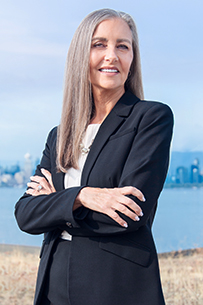 Pauline stands in a relaxed three-quarter pose with her arms crossed, smiling, dressed in a grey skirt suit and white shirt on a light grey background.