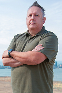 Gary stands in a relaxed three-quarter pose, smiling, wearing a dark blue blazer over a light blue collared shirt and dark jeans against a light grey background.