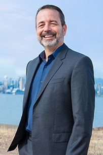 Jason stands in a relaxed three-quarter pose, smiling, dressed in a dark grey suit and light blue shirt on a light grey background.