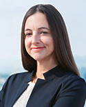 Kelly smiles at the camera, wearing a medium grey blazer against a light grey background.