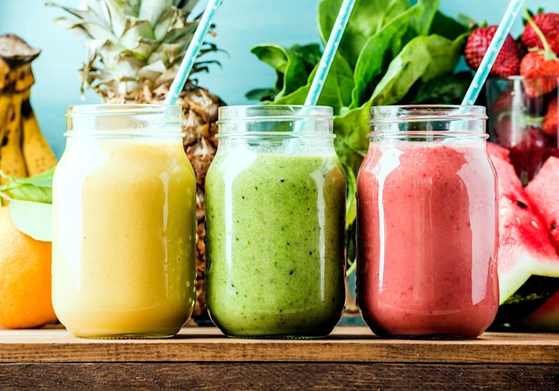 Three jars of colourful smoothies on a table with fruit in the background