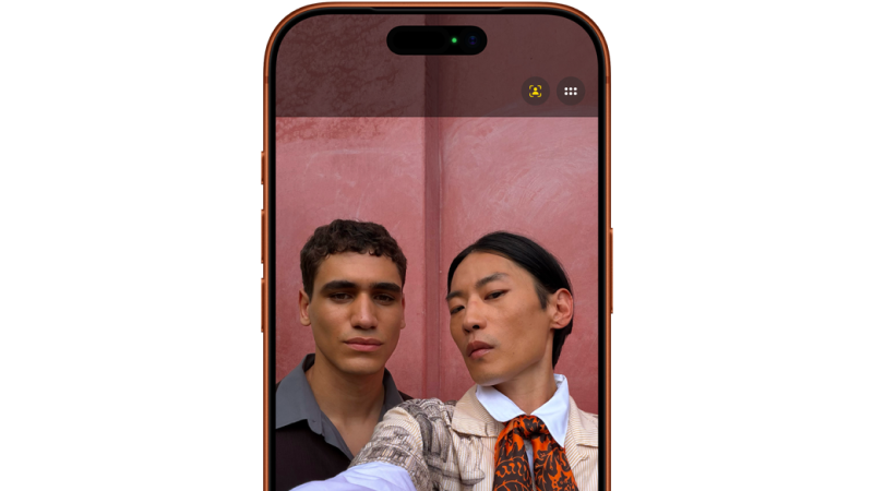 Två personer tar en selfie mot en rosa vägg, visas på en smartphone-skärm med orange ram.