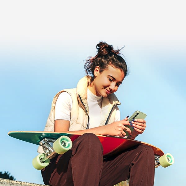 Tjej sitter med en skateboard i knät i dagsljus och använder Samsung Galaxy A54