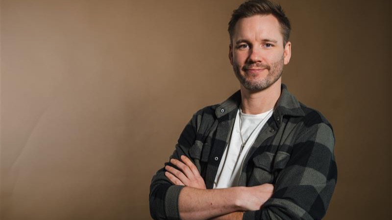 En person med skägg i rutig skjorta och vit t-shirt står med armarna i kors mot en beige bakgrund.