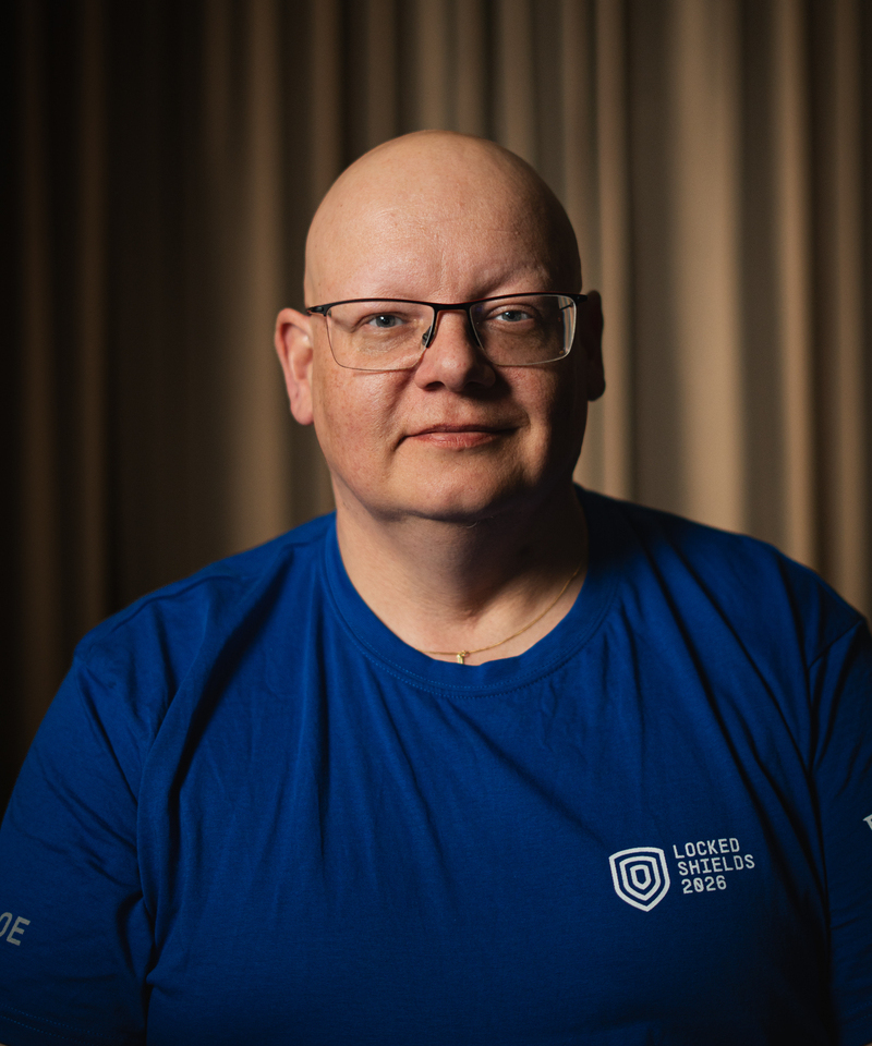 Man med glasögon klädd i blå t-shirt med text "Locked Shields 2026". 