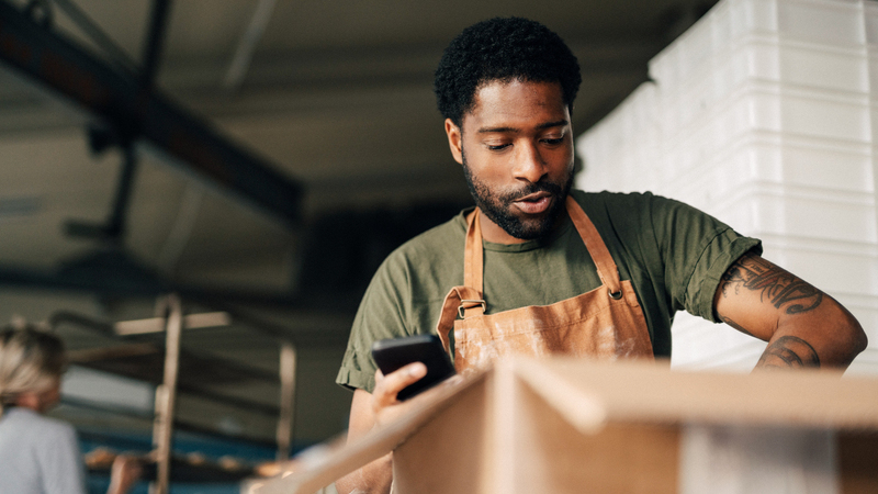 En man med mobil i ett restaurangkök samt en kvinna i bakgrunden. 