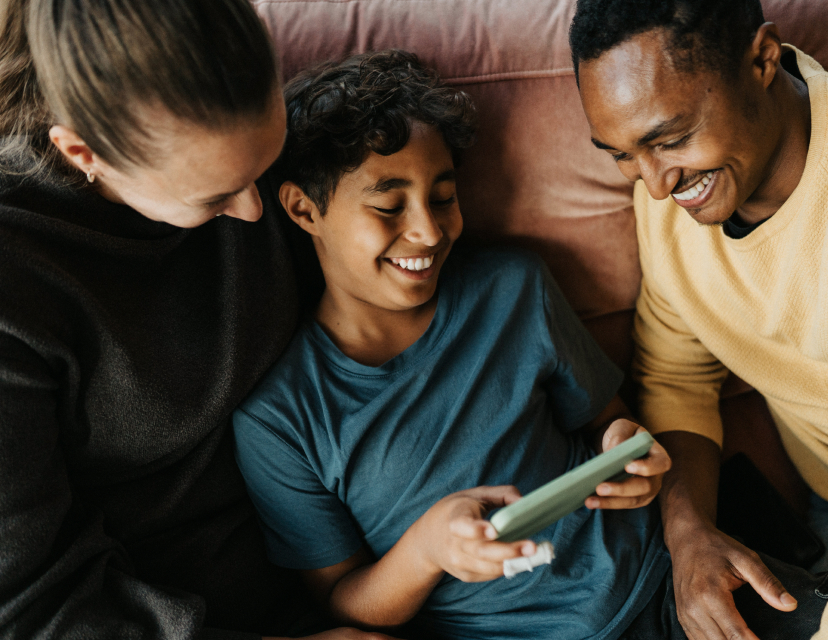 Tre personer sitter tillsammans i en soffa och tittar glatt på en mobiltelefon som barnet i mitten håller.
