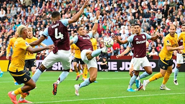 En intensiv fotbollsscen där spelare från Aston Villa i vinröda tröjor och Wolverhampton Wanderers i gula tröjor kämpar om bollen inne i straffområdet under en match. En Aston Villa-spelare försöker sparka bollen medan andra spelare från båda lagen är nära. Publiken i bakgrunden följer matchen noggrant.