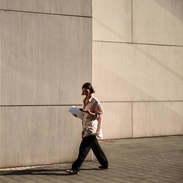 En kvinna går förbi en grå betongvägg i starkt solljus. Hon är klädd i skjorta och byxor och håller i en bunt papper samt en mobiltelefon. 