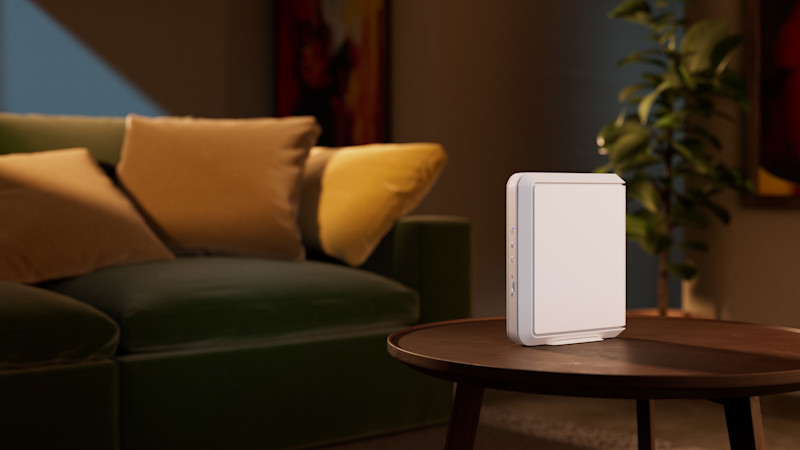 Vit router på träbord bredvid grön soffa med gula kuddar i ett hemtrevligt vardagsrum med krukväxt i bakgrunden.