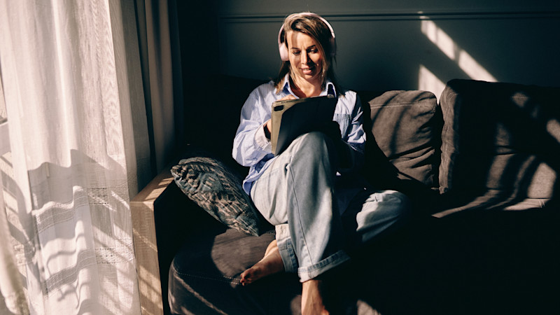 Person med hörlurar sitter avslappnat i soffa nära fönster med solljus, tittar på surfplatta och ler.