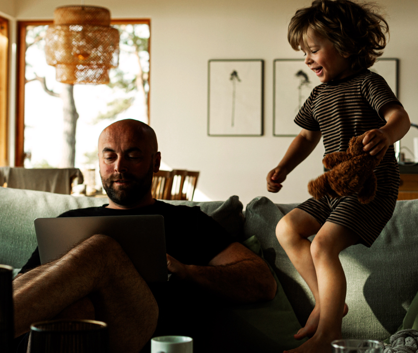 Glad pojke med nallebjörn hoppar i soffa medan vuxen arbetar på laptop i ett ljust vardagsrum med rotting-lampa.