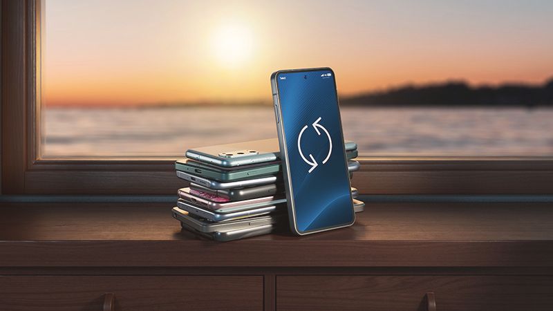 En hög med mobiltelefoner ligger på ett fönsterbräde. En telefon med återvinningssymbol på skärmen står lutad mot dem. Utanför fönstret syns vatten och solnedgång. 