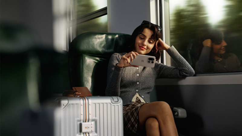 En kvinna sitter på ett tåg i rörelse och tittar på en mobilskärm. Bredvid henne står en silverfärgad resväska och utanför skymtar träd och himmel. 