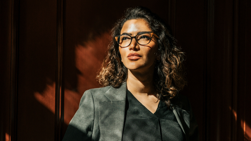 Kvinna med brunt hår och glasögon, klädd i grå kavaj, står lutad mot en röd husvägg. 