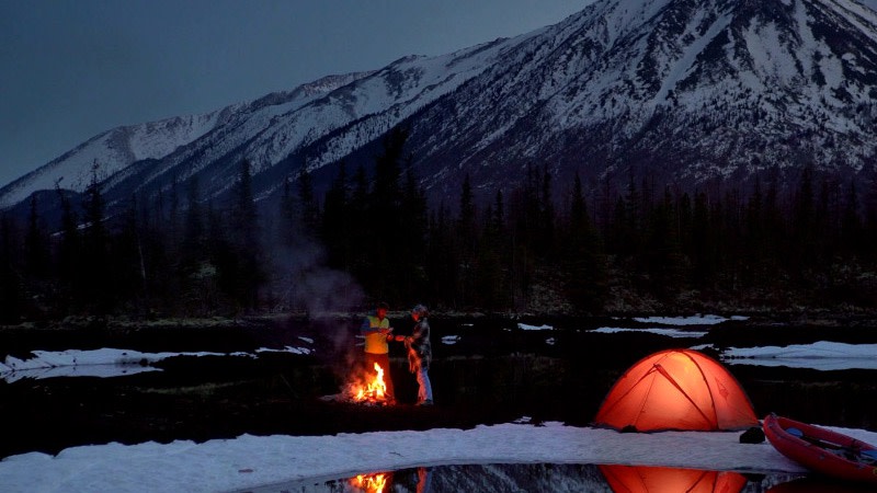 Campingplats i skymningen med upplyst orange tält och lägereld framför snötäckt berg och mörk barrskog.