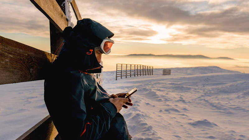 Person i skidkläder tittar på mobiltelefon vid solnedgång över snötäckt landskap med staket i bakgrunden.