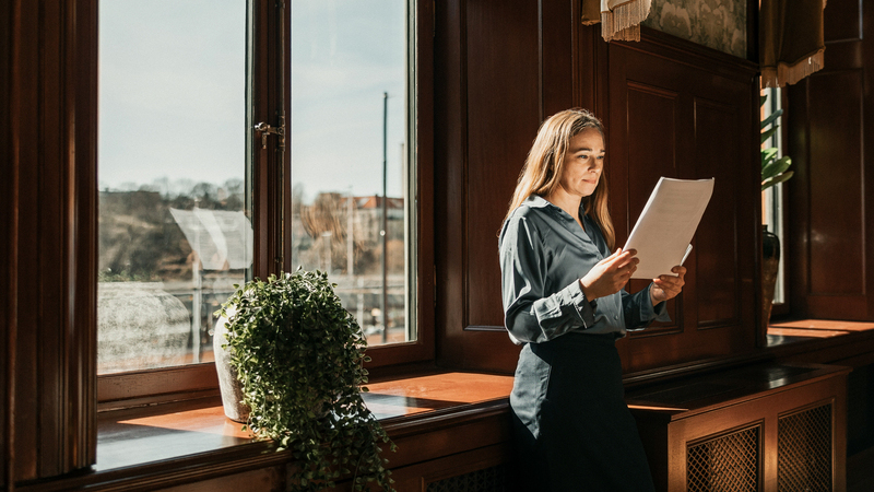 Kvinna står med ryggen mot ett fönster på ett kontor. Hon läser på ett papper och befinner sig på ett kontor. 