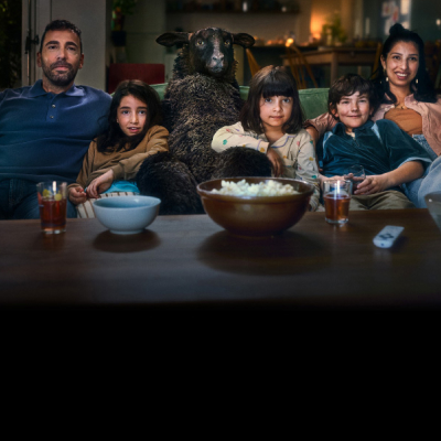 En familj sitter i en soffa med en stor svart får-liknande figur i mitten, tittar på TV med popcorn och drycker framför sig.
