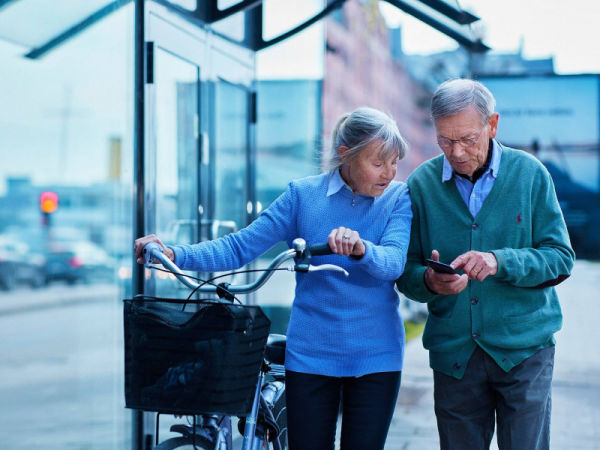 Ett äldre par i stadsmiljö. Kvinnan leder en cykel medan mannen visar henne något på sin mobil.