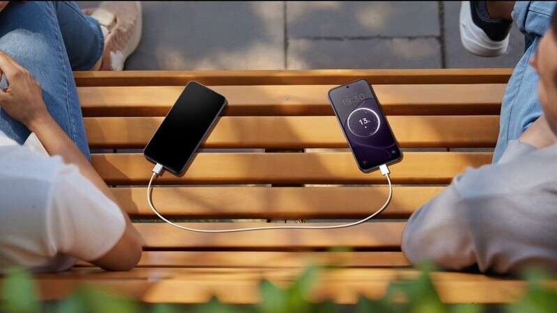 Två personer sitter på en bänk. Mellan dem ligger två mobiler som delar batteri genom en vit kabel. 