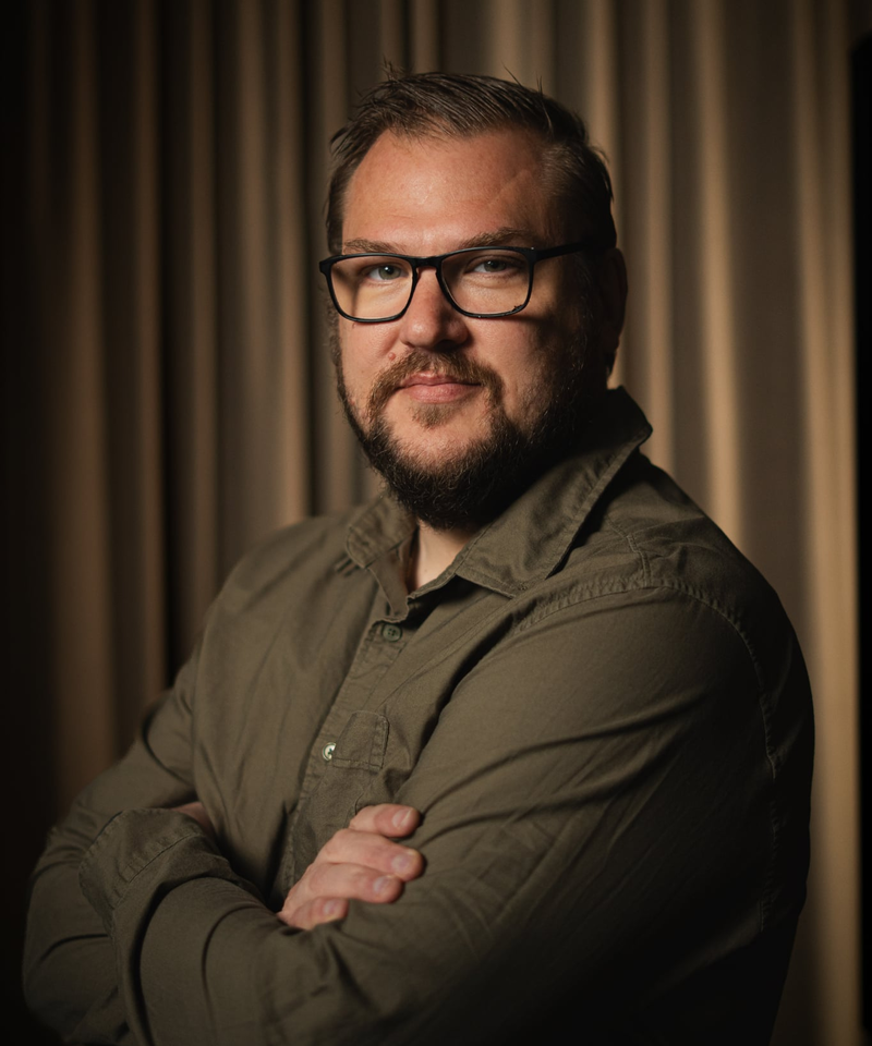 En man med mellanblont hår och skägg klädd i mörkgrön skjorta mot en beige bakgrund. 