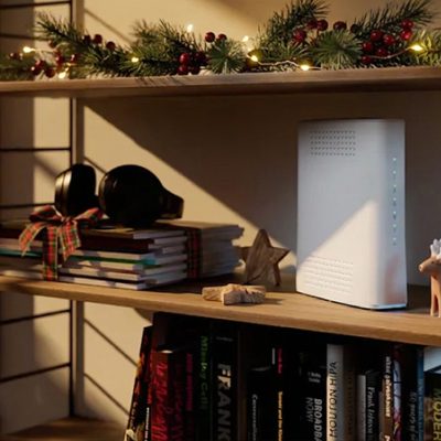 En bokhylla med en vit router, böcker och juldekoration med gröna kvistar och röda bär överst, upplyst av små ljus.