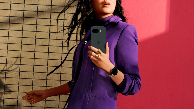 Kvinna med långt svart hår som håller i en svart smartphone av modell Google Pixel 10a. Hon är klädd i en lila klänning och står framför en röd vägg. 