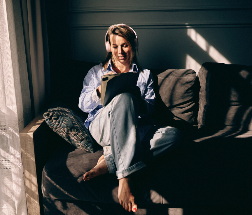 Person med hörlurar sitter avslappnat i en soffa och tittar på en surfplatta, solljus strömmar in genom fönstret.