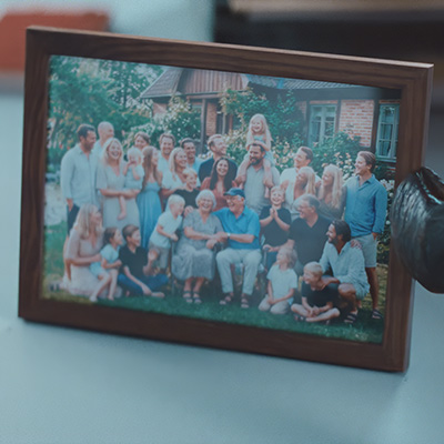 En inramad familjefoto med många generationer som poserar tillsammans utomhus framför ett hus med grön trädgård.