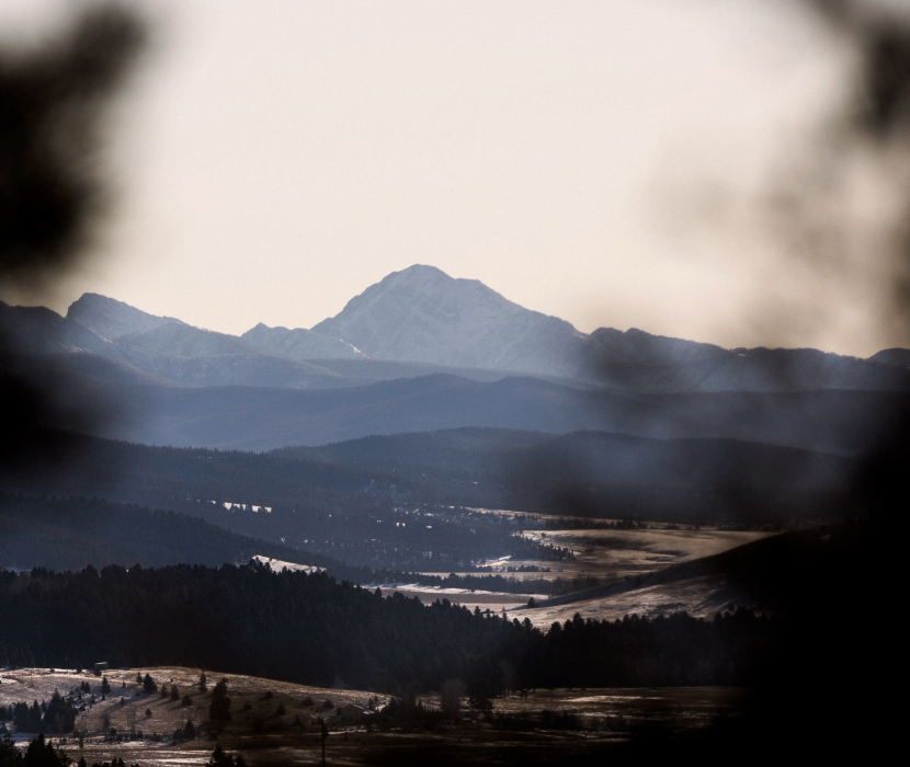 Dimmigt bergslandskap med blåaktiga bergskedjor i bakgrunden och dalar med skogsområden i förgrunden under en disig himmel.