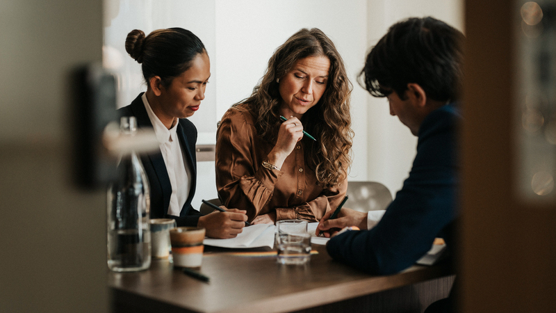 Två kvinnor och en man sitter vid ett bord med papper och kaffekoppar framför sig. 
