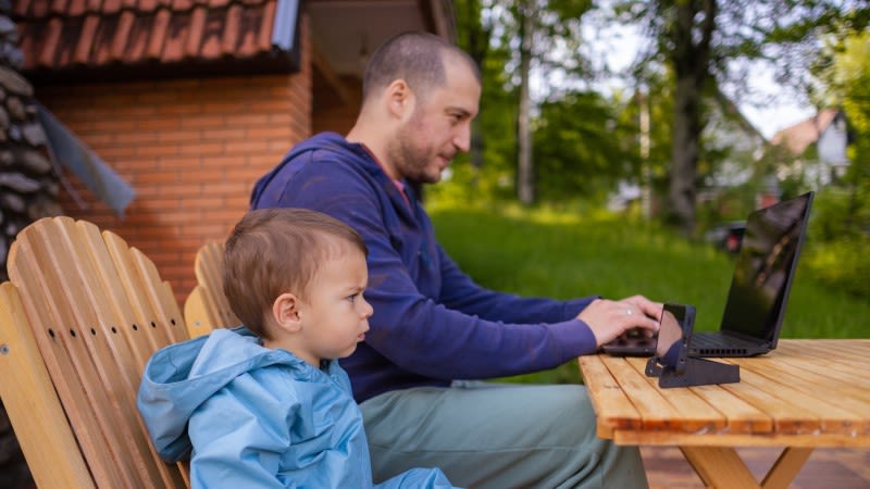 En man sitter på en uteplats och jobbar på sin dator. Bredvid honom sitter en liten pojke och tittar på något på en mobil. Pojken är djupt koncentrerad.