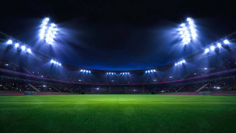 En upplyst fotbollsstadion på natten med grönt gräs i förgrunden och starka strålkastare mot mörk himmel.
