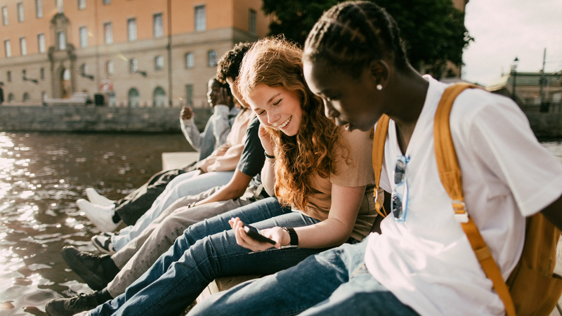 En grupp ungdomar sitter på en bänk och tittar på mobiltelefoner. 