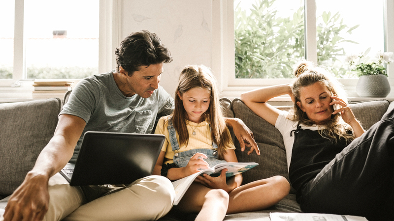 En man och en flicka sitter i en soffa med en laptop och en skolbok. Bredvid dem sitter en kvinna och pratar i mobiltelefon. 